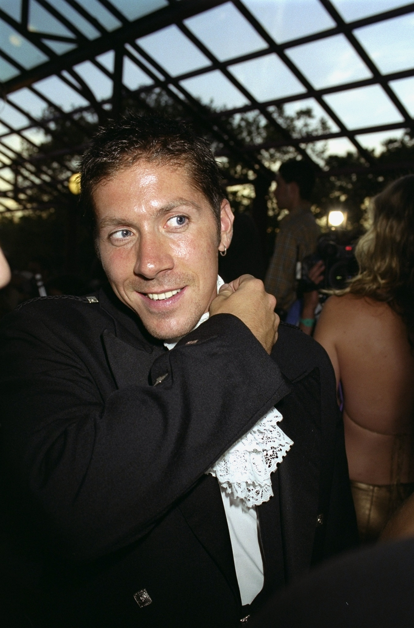 A person at an event, wearing a formal attire with a lace collar, looks to the side while adjusting their collar. The background shows other attendees