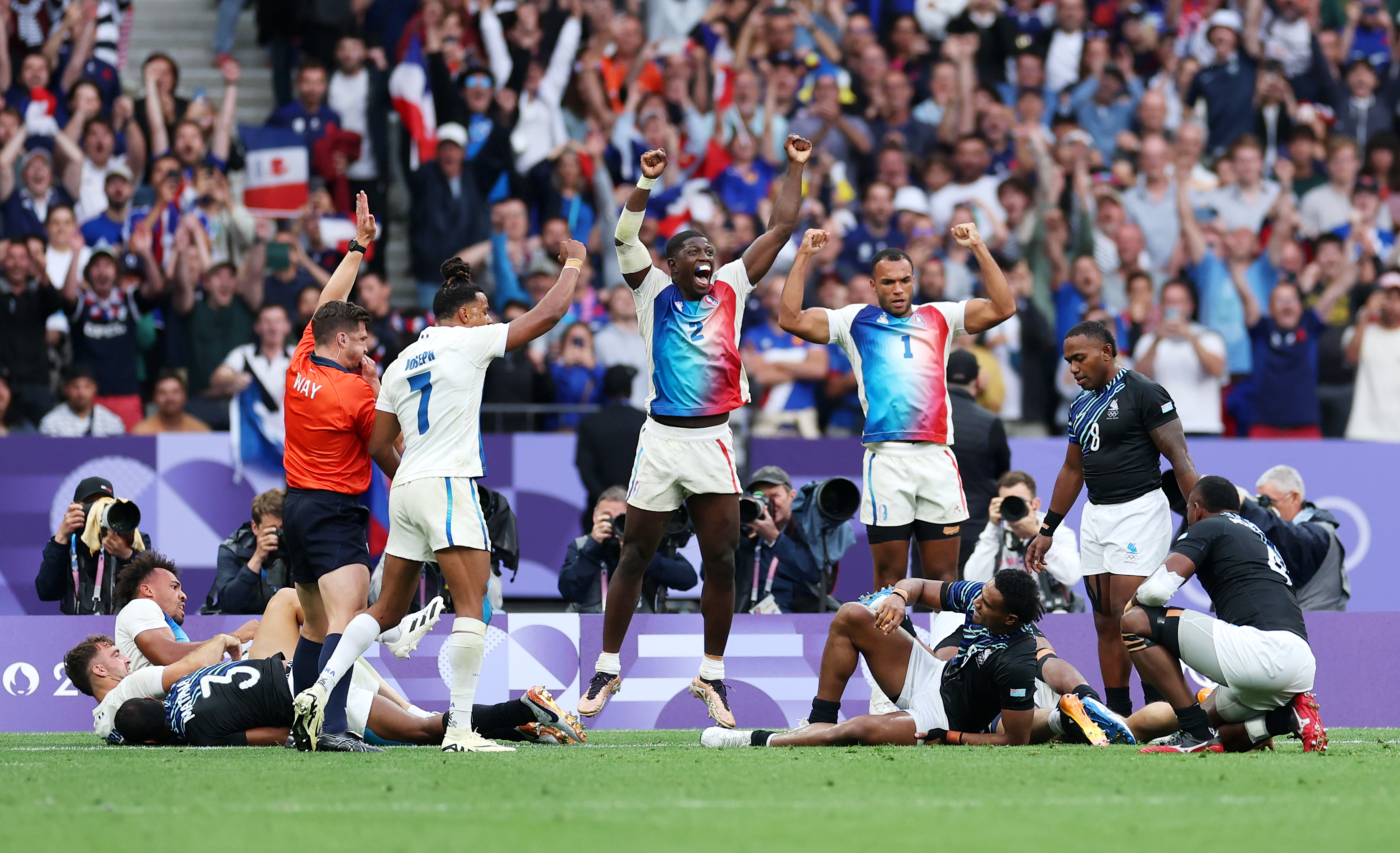 Rugby players celebrate on the field after scoring a try during a match, with one referee signaling and the crowd cheering in the background