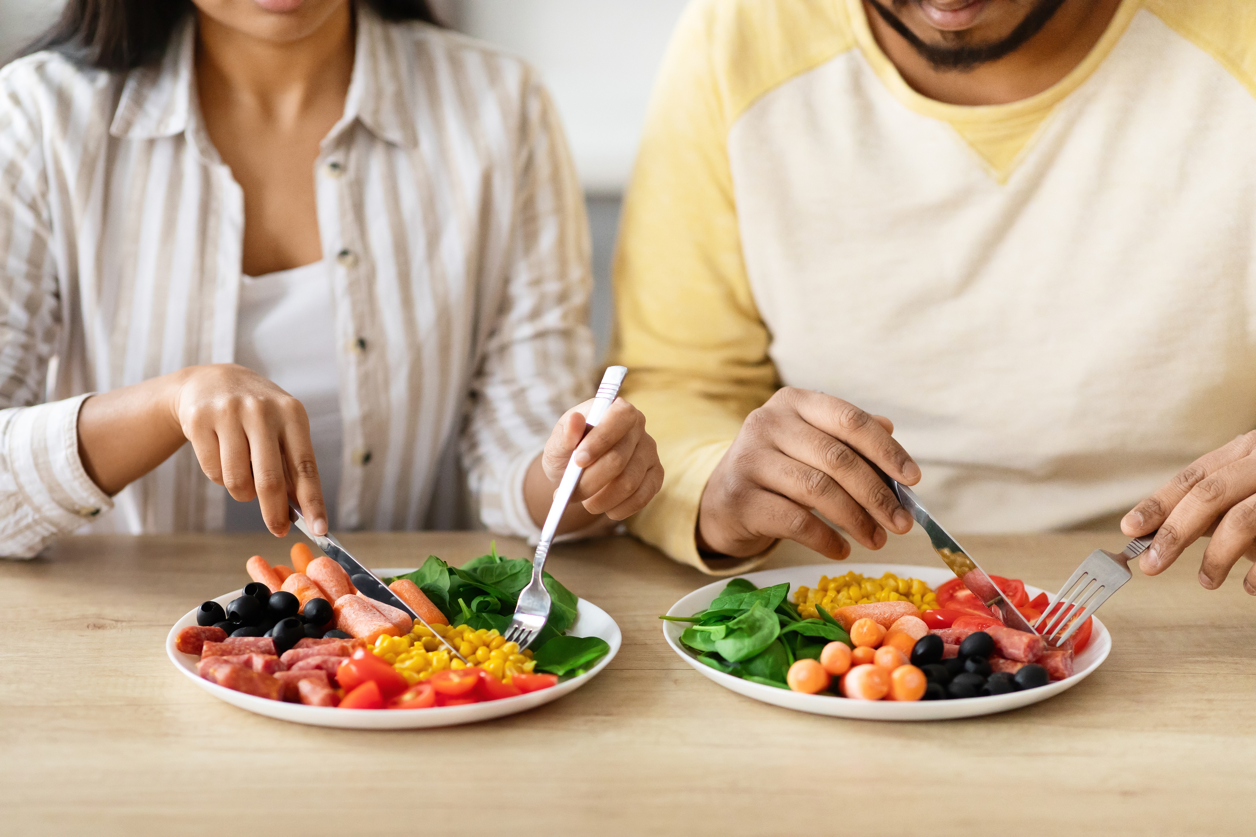 Two unidentified people dining on plates of assorted fresh vegetables, olives, corn, and meat slices