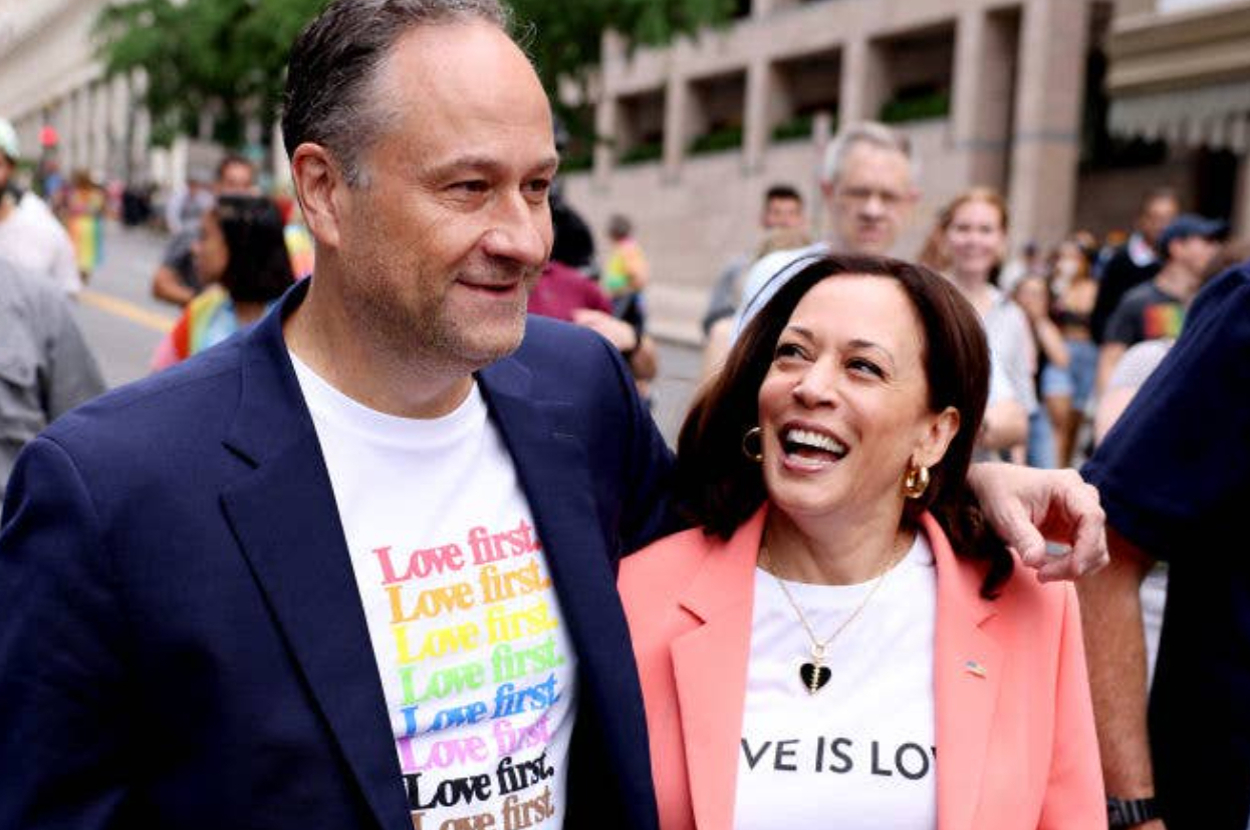 Doug Emhoff and Kamala Harris walk together in a pride parade. They are smiling and wearing shirts with pride-themed messages