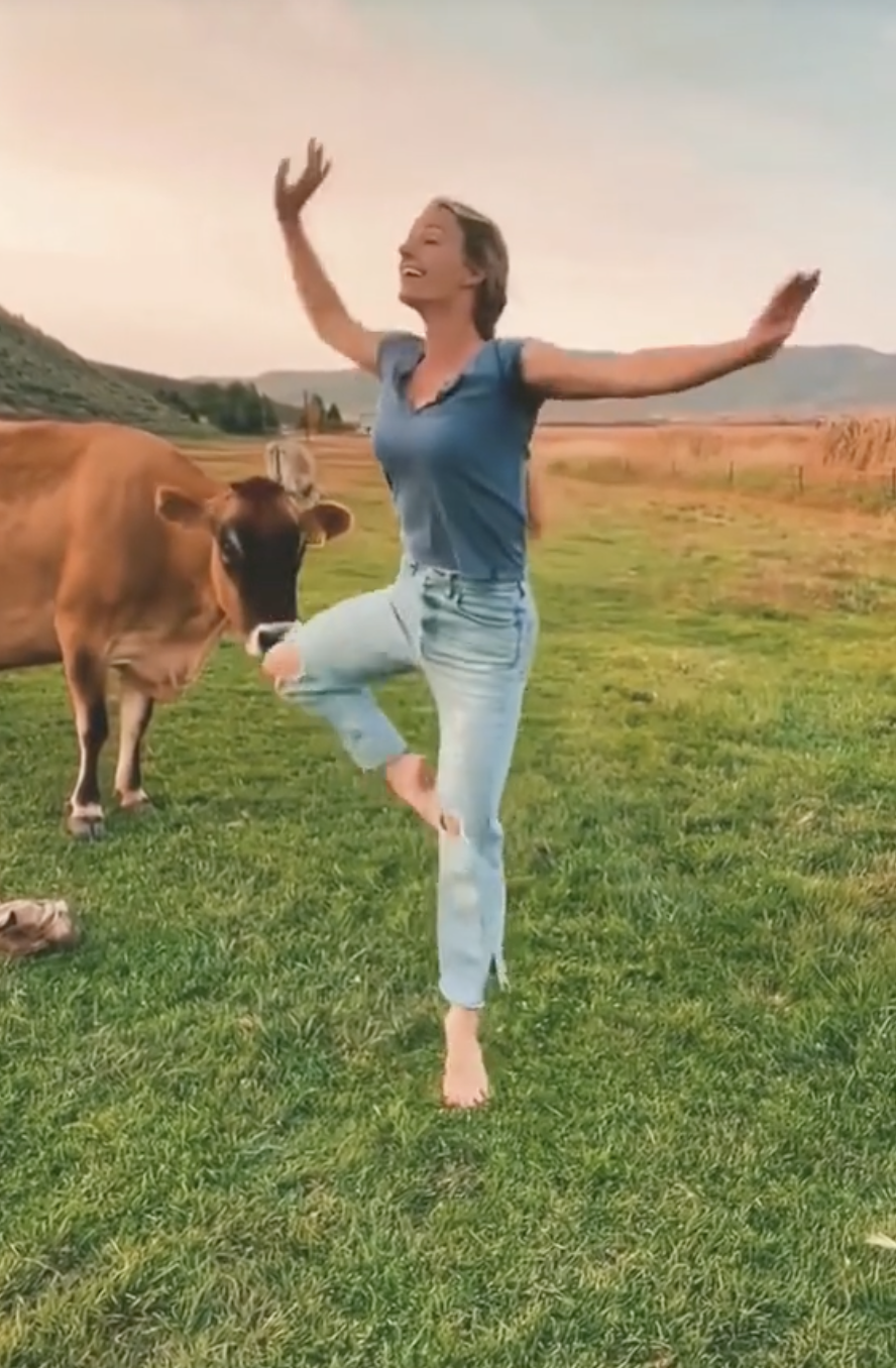 A woman joyfully dances barefoot on grass beside a nearby cow in a rural setting