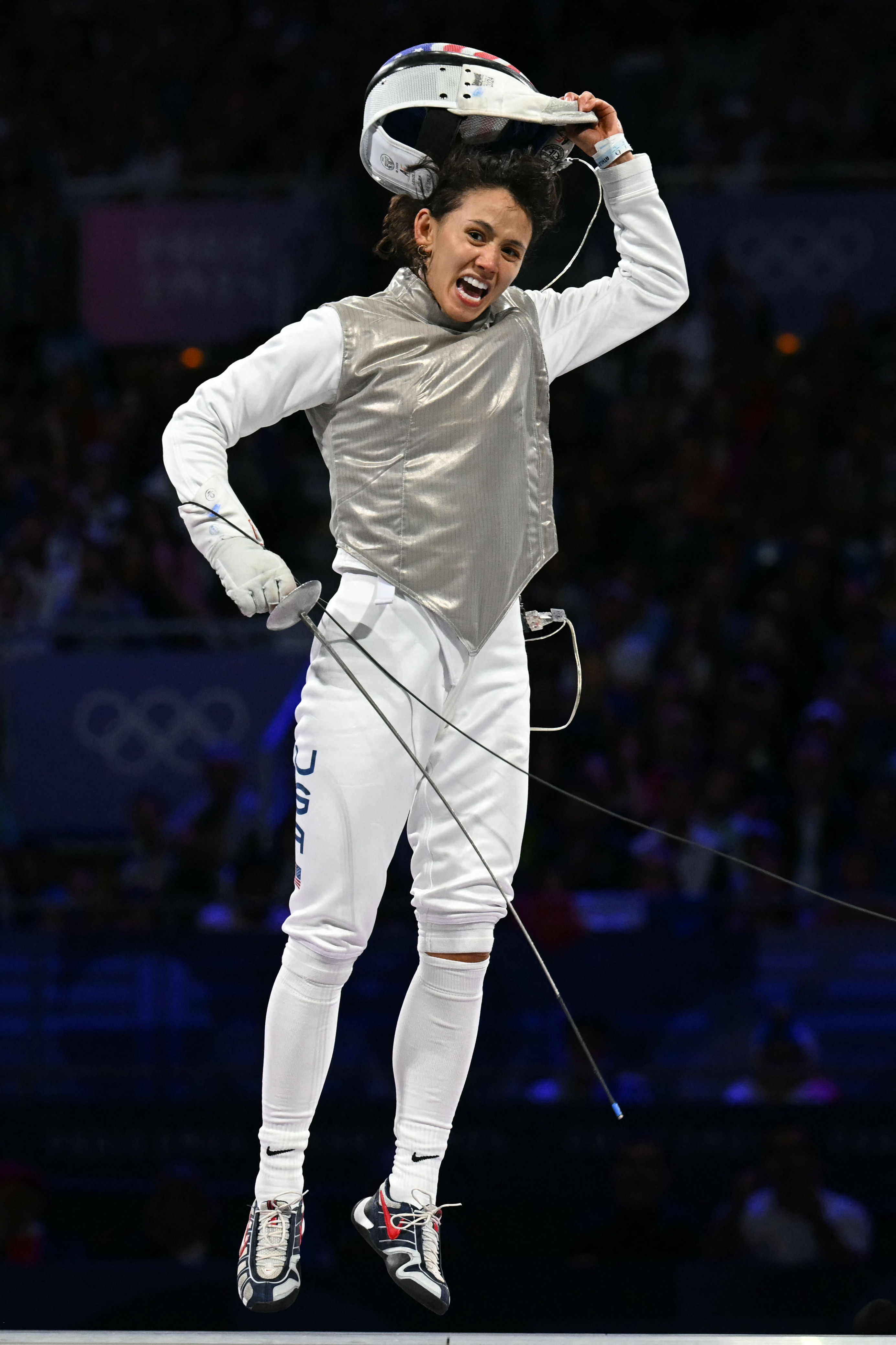 Olympic fencer Ibtihaj Muhammad jumps joyfully, holding her helmet aloft after a match, dressed in full fencing attire