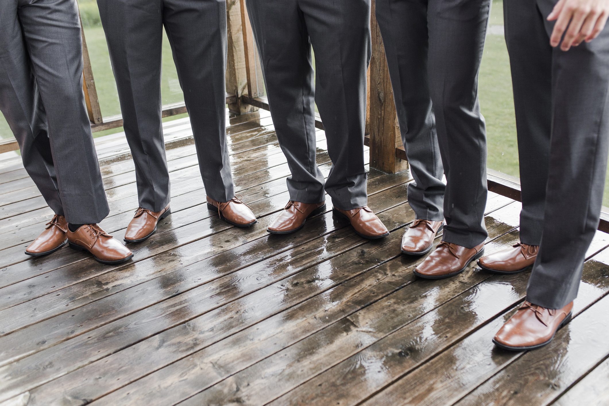 Five people wearing grey suits and brown dress shoes are standing on a wet wooden deck. Their faces are not shown
