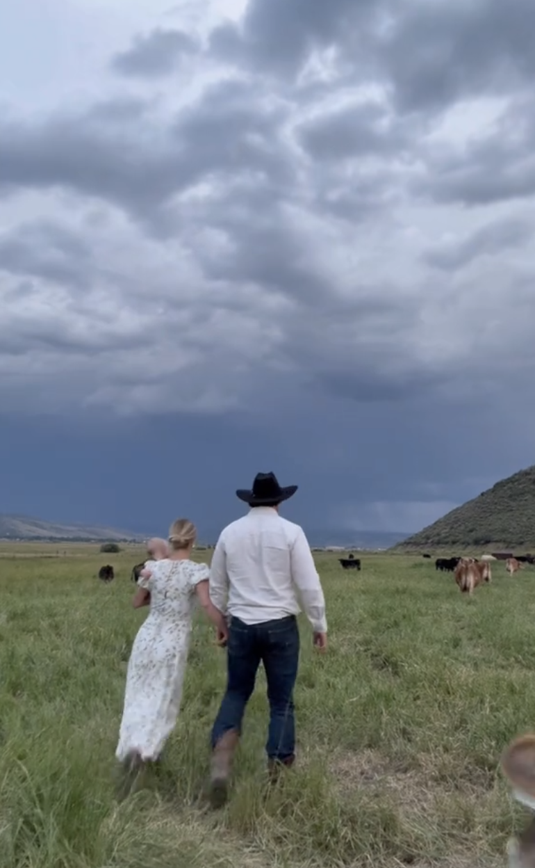 A woman in a floral dress and a man in a cowboy hat walk hand in hand through a grassy field, with cows grazing nearby under a cloudy sky