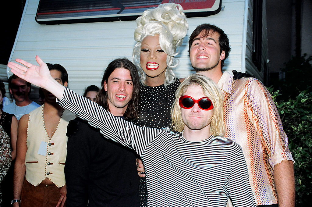 Dave Grohl, RuPaul, and Kurt Cobain posing together with another unidentified person, dressed in '90s fashion, smiling and happy at an outdoor event
