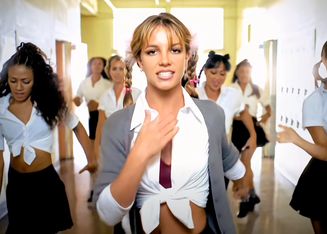 Britney Spears, in a tied white shirt and gray cardigan, dances in a school hallway with backup dancers in similar outfits from the "Baby One More Time" music video
