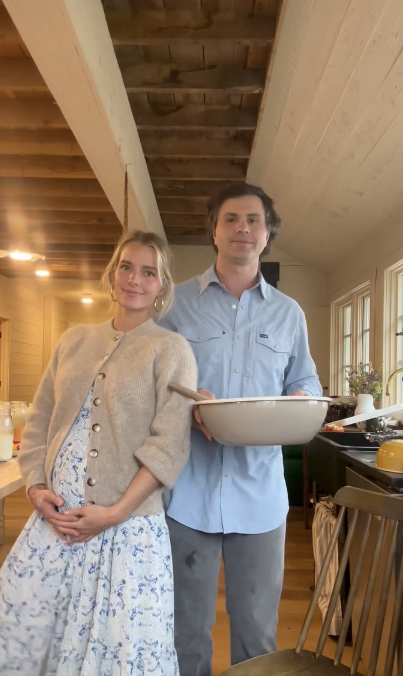 Hannah and Daniel standing in a kitchen, smiling. Hannah is wearing a floral dress and cardigan, while Daniel is in a shirt holding a bowl with a spoon