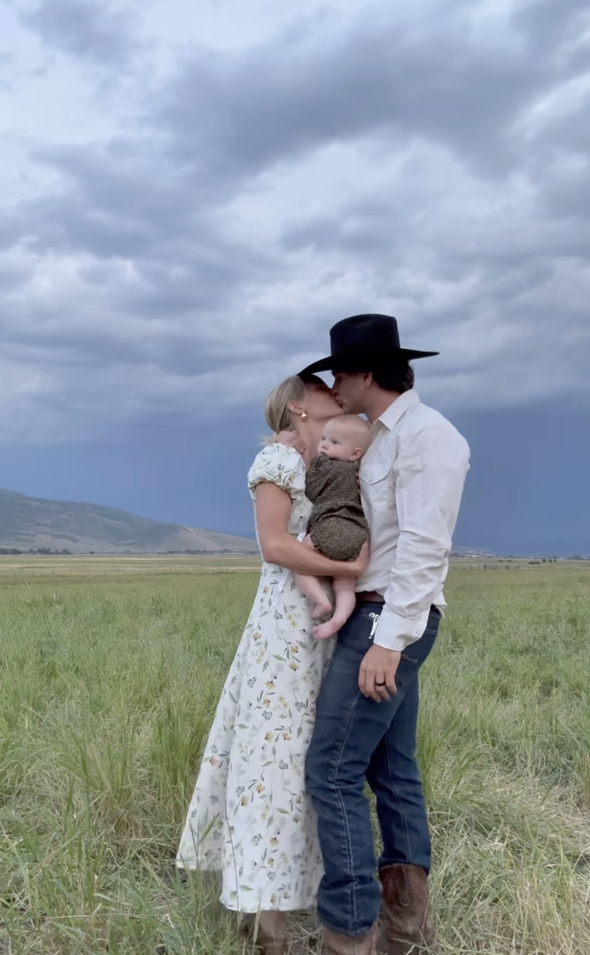 A couple, with the man wearing a cowboy hat, shares a kiss while holding a baby in a grassy field. Dark clouds loom in the sky
