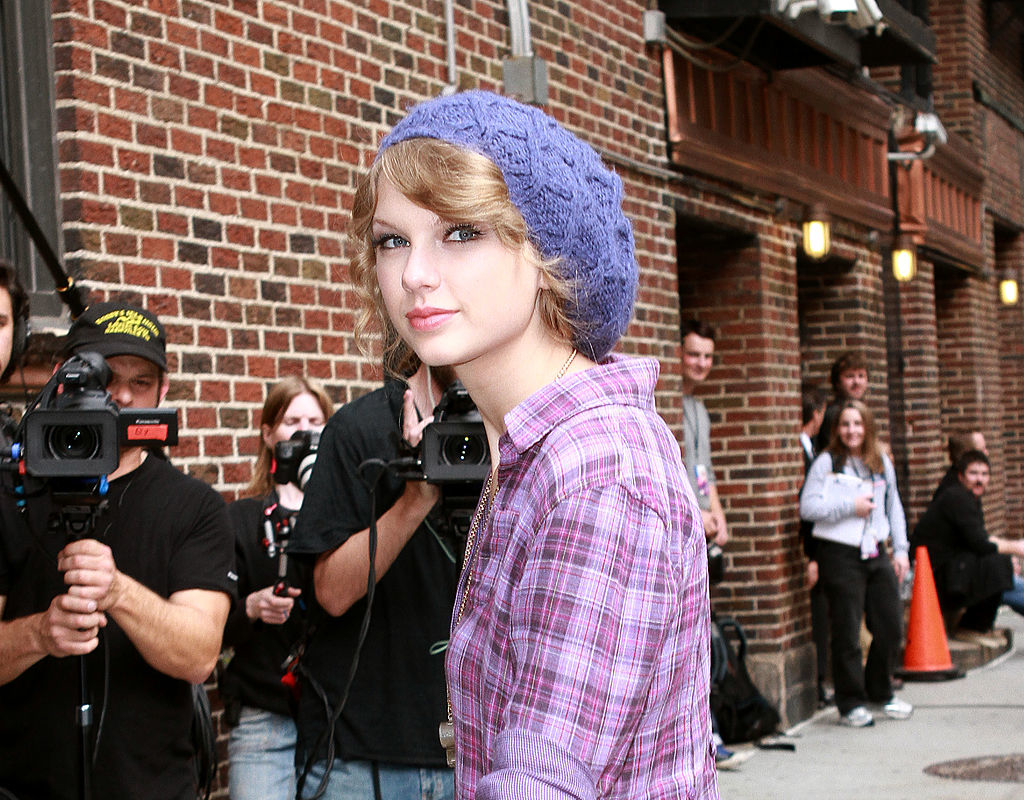 Taylor Swift outdoors wearing a casual plaid shirt and a knitted beanie, with paparazzi and fans in the background