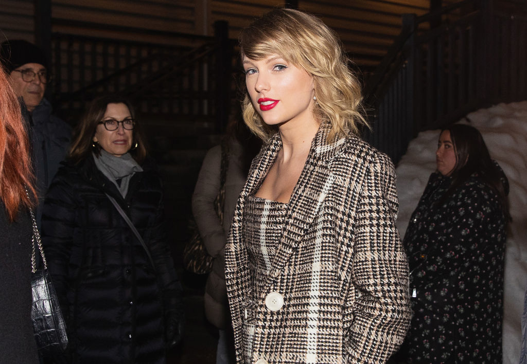 Taylor Swift in a chic houndstooth coat at an outdoor evening event, surrounded by people