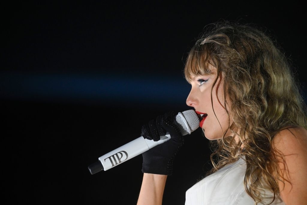 Taylor Swift performs on stage, holding a microphone and wearing a black glove and white outfit