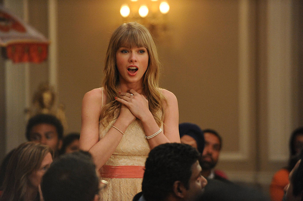 Taylor Swift standing with hands clasped to her chest, looking surprised and smiling, in a room with several other people in the background