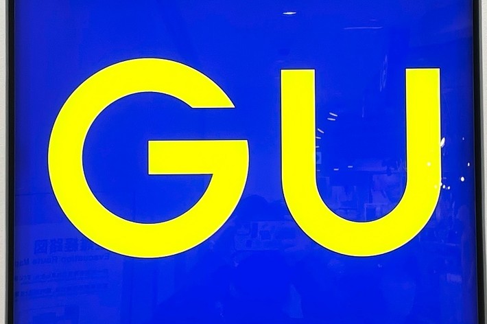 A blue square sign with yellow letters &ldquo;GU&rdquo; in the center, mounted on a white wall