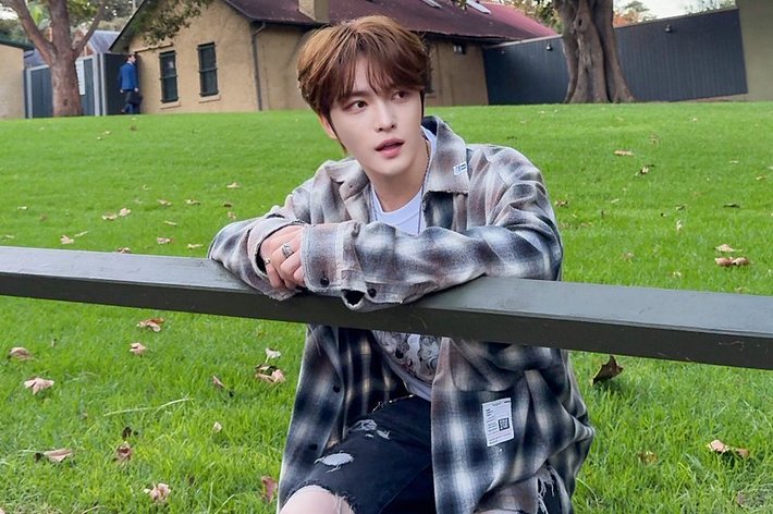 芝生にいる人物が木製の柵に手をかけてしゃがんでいます。背景には古い建物が見えます。