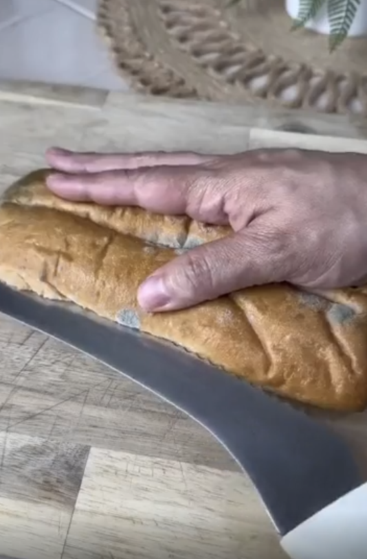 A hand holding a loaf of bread on a cutting board, about to slice it with a large knife