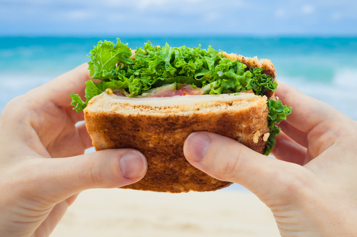 A close-up of hands holding a sandwich filled with fresh vegetables, with the ocean and beach in the background