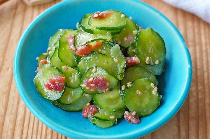 青いお皿に盛られた、胡瓜と梅干しの和え物。お箸と布が背景に見える。