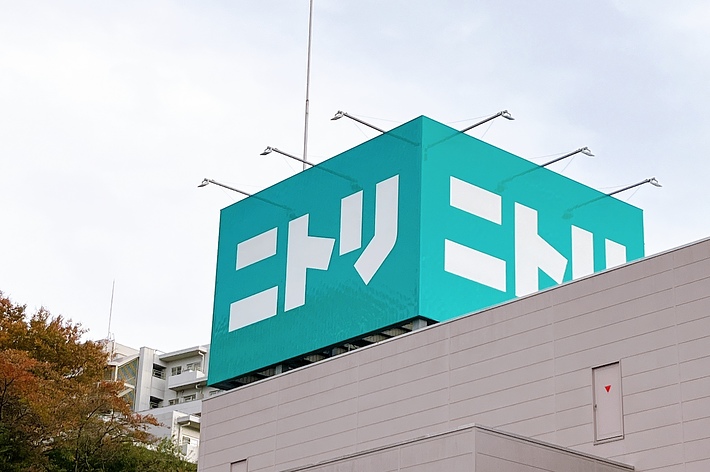 ニトリの広告看板が建物の屋上に設置されている。背景に秋の木々と建物が見える。