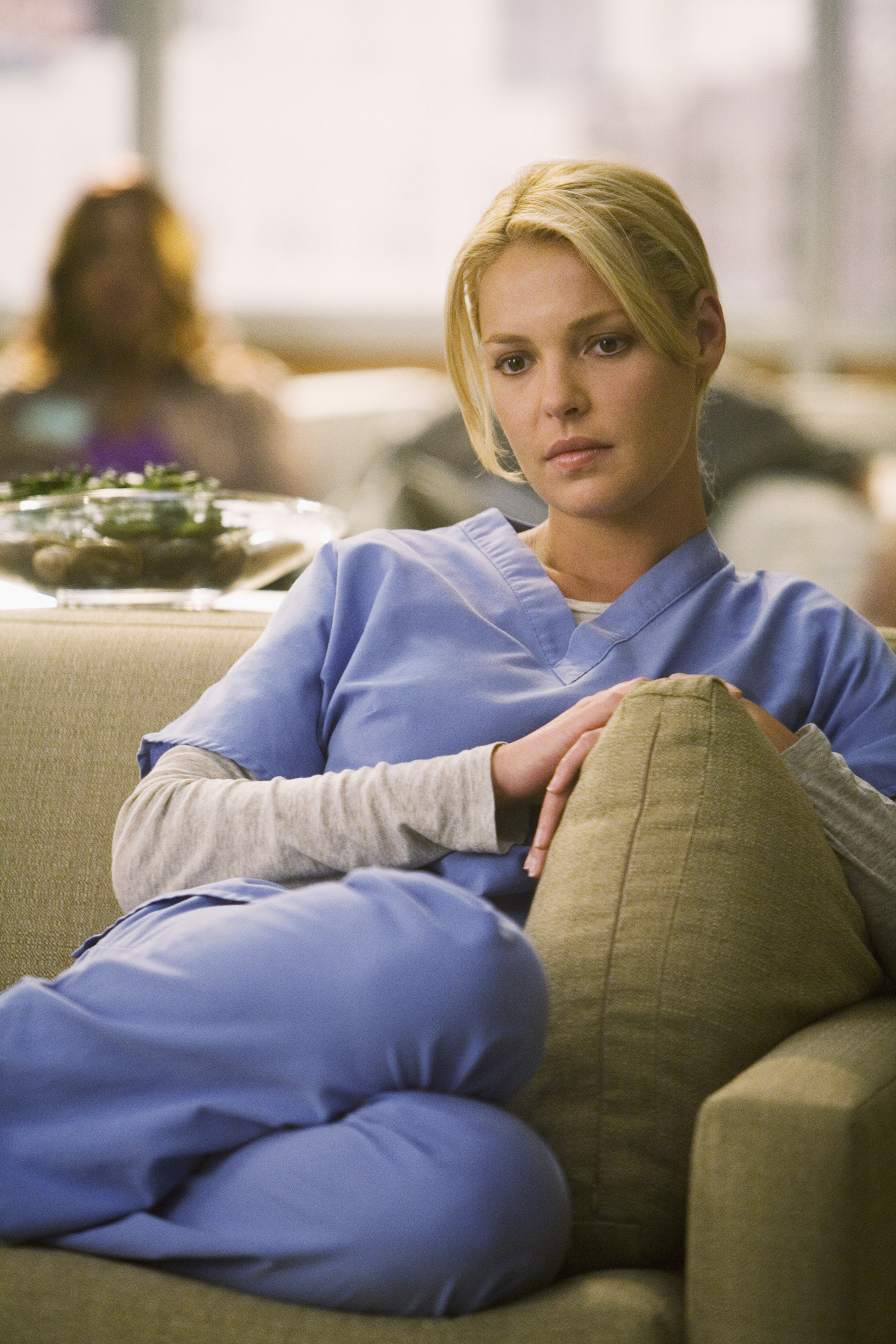 Katherine Heigl, wearing hospital scrubs, sits on a couch looking contemplative