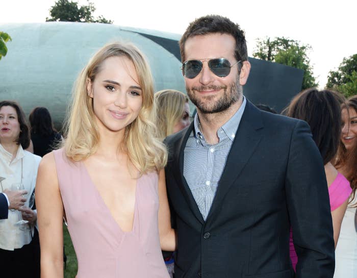 Suki Waterhouse in a sleeveless dress and Bradley Cooper in a suit and sunglasses, smile at an outdoor event