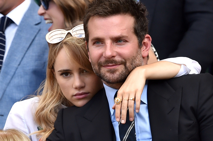 Bradley Cooper in a black suit and Suki Waterhouse wearing sunglasses sit closely at a public event, surrounded by other attendees