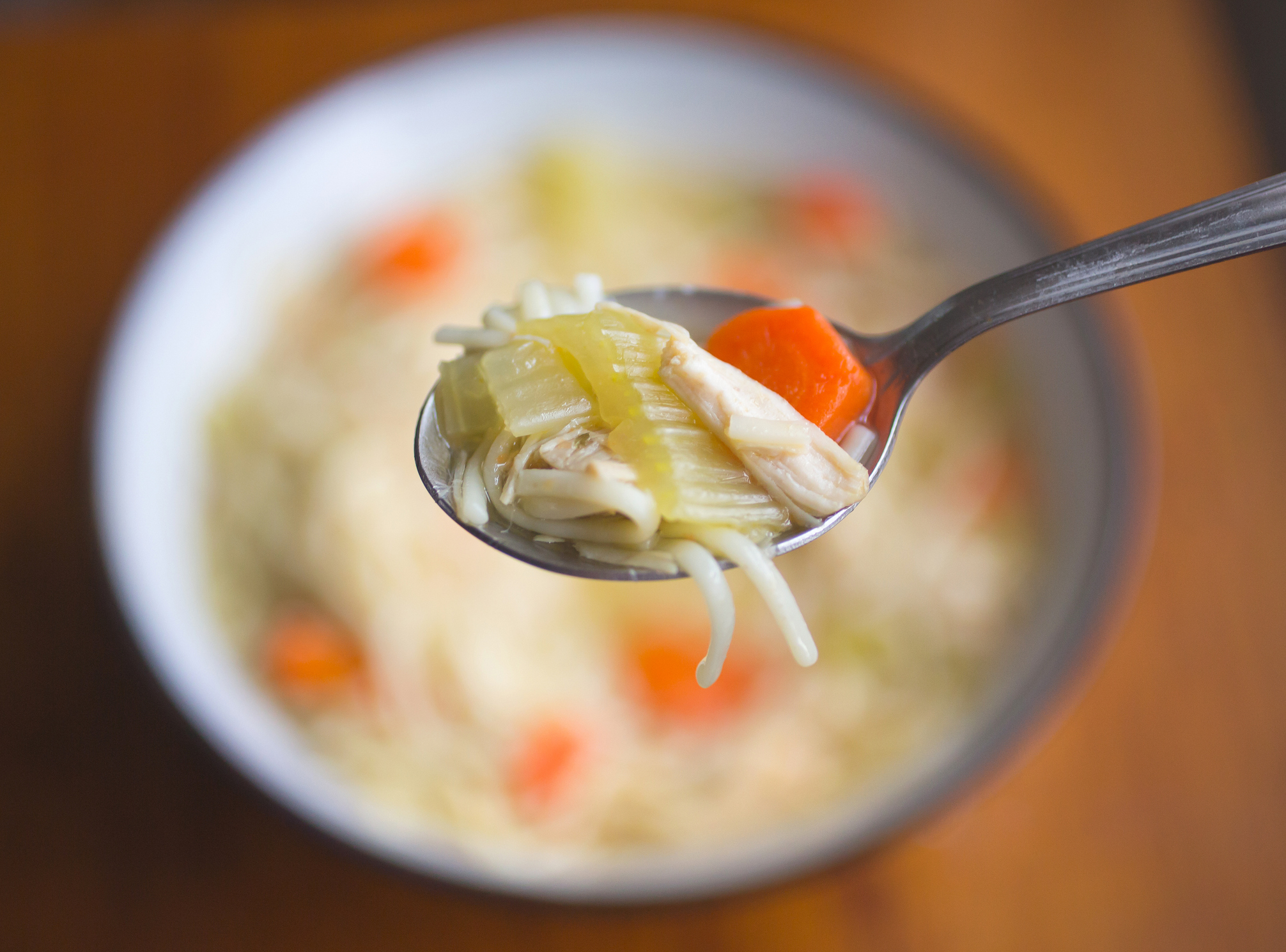 Spoonful of chicken noodle soup with carrots and celery, held over a bowl of soup in the background