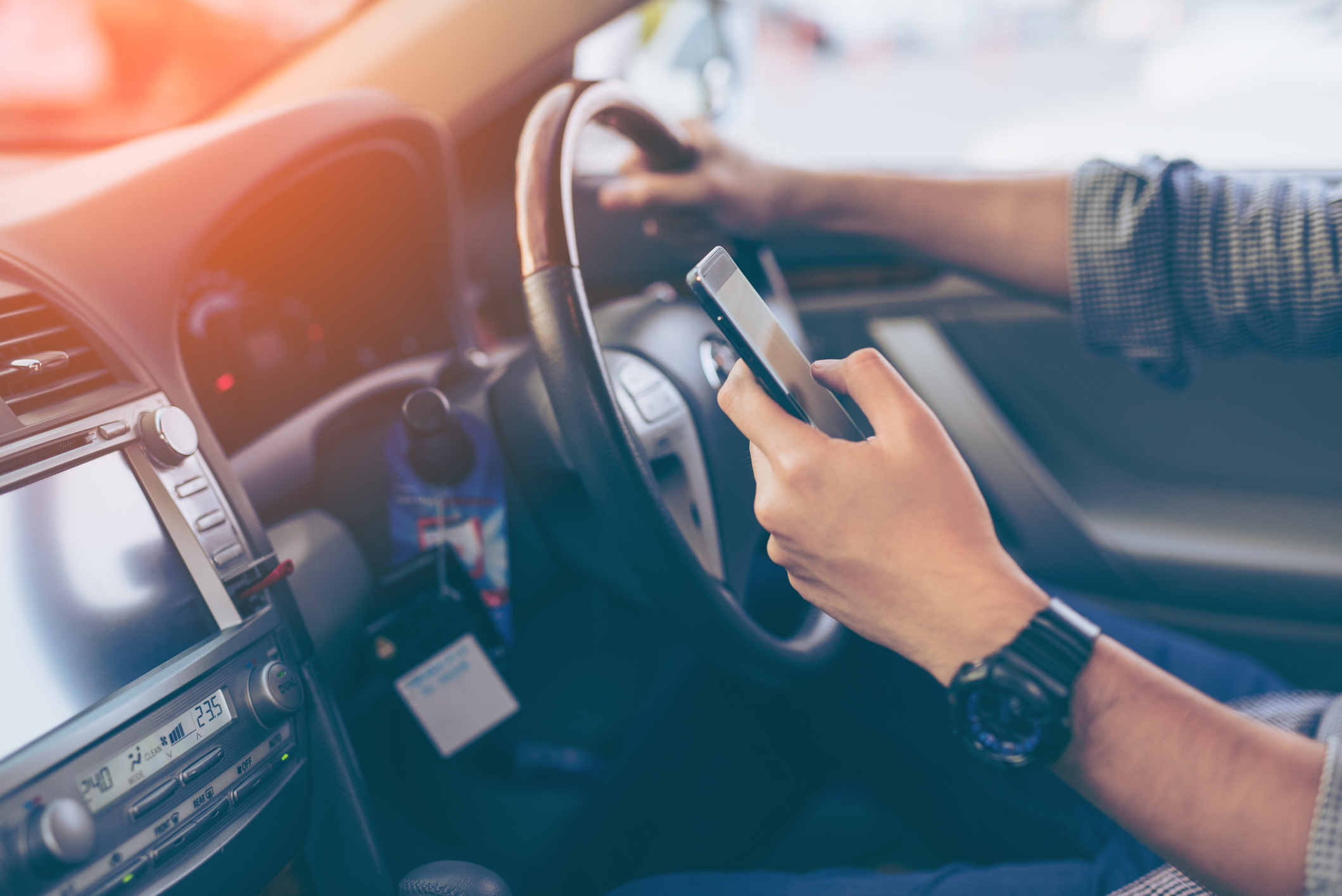 A person driving while holding a phone in one hand, their other hand on the steering wheel. Focus on the phone use while driving