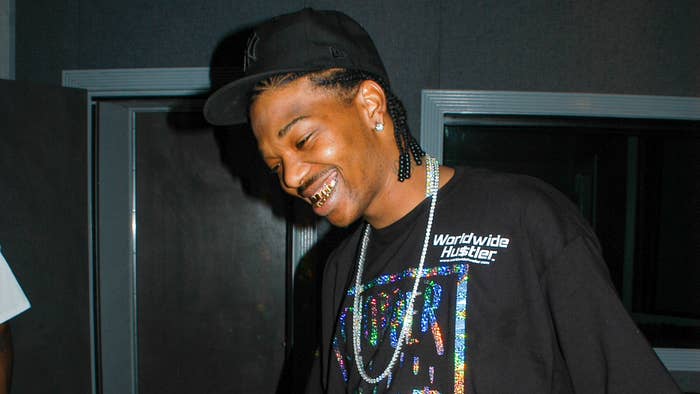 A smiling man with braids and gold teeth wears a black cap and a T-shirt that reads &quot;Worldwide Hustler&quot; in a recording studio