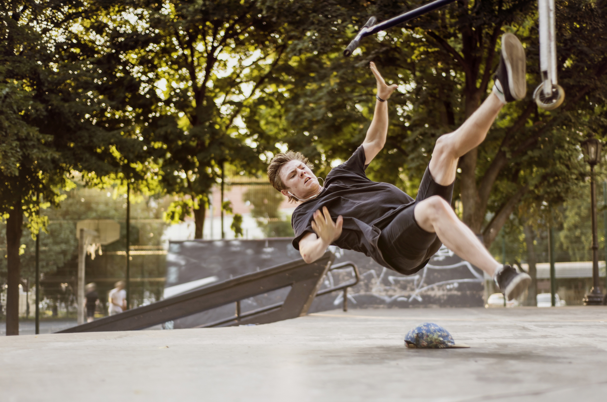 Person mid-air after falling off a scooter at a skate park. A helmet is on the ground. Trees and people in the background