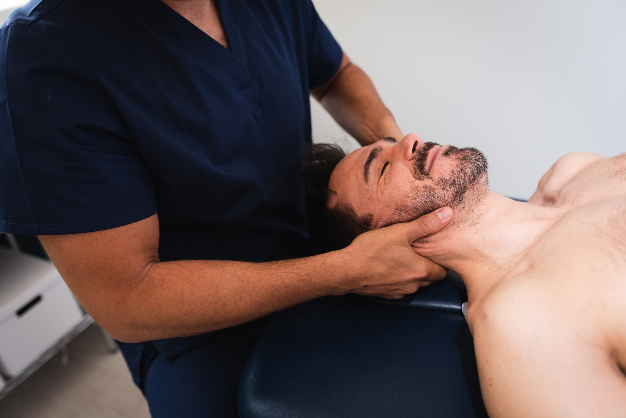 A person in a navy medical uniform performs a neck adjustment on a man lying shirtless on a treatment table