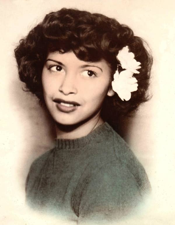 A young woman with curly hair and a flower in her hair, wearing a knitted top, poses for a portrait