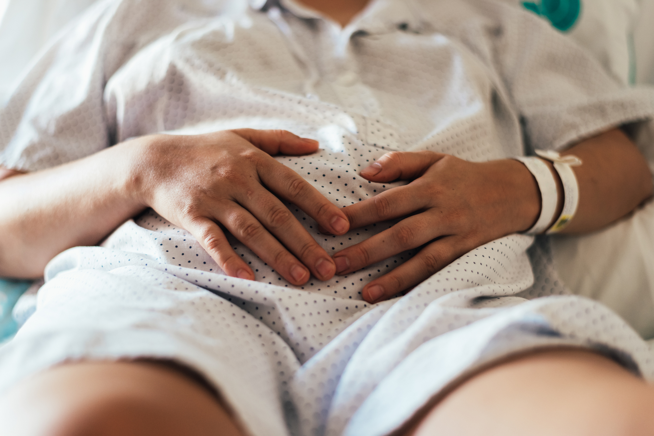 A close-up shot of a person lying down, wearing a hospital gown, with their hands resting on their pregnant belly