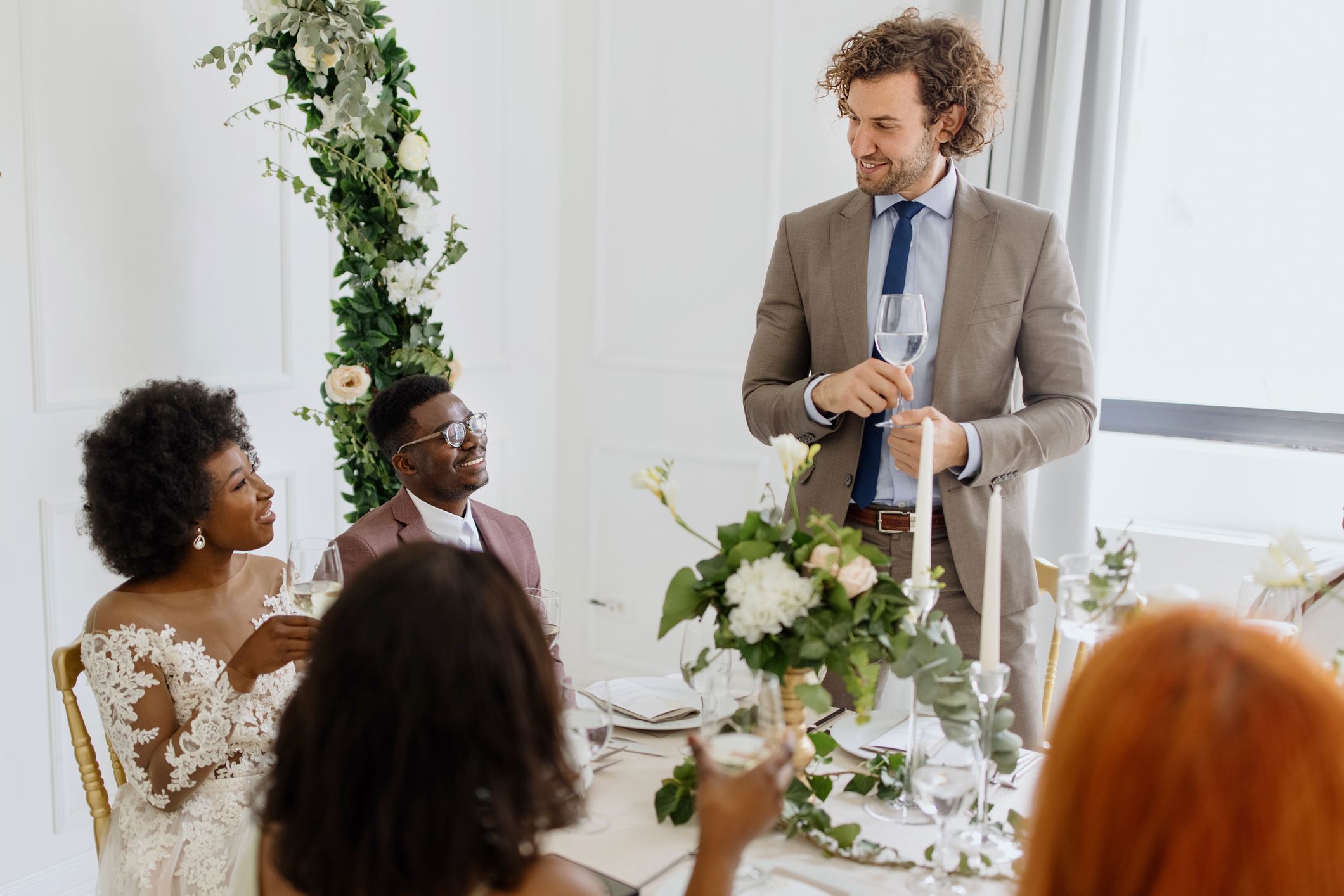 A wedding toast is given by a man in a light suit while a woman in an off-shoulder wedding dress and a man in a dark suit are seated, smiling at him
