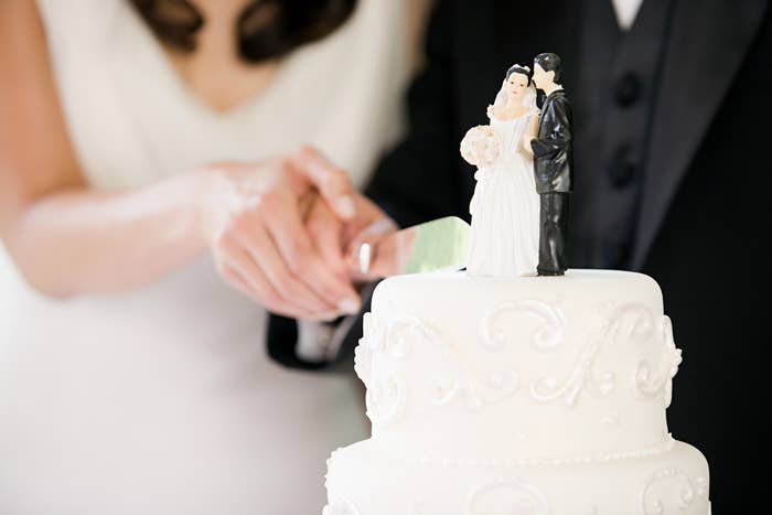A bride and groom cut a wedding cake adorned with a figurine of a couple in wedding attire. Their faces are not visible