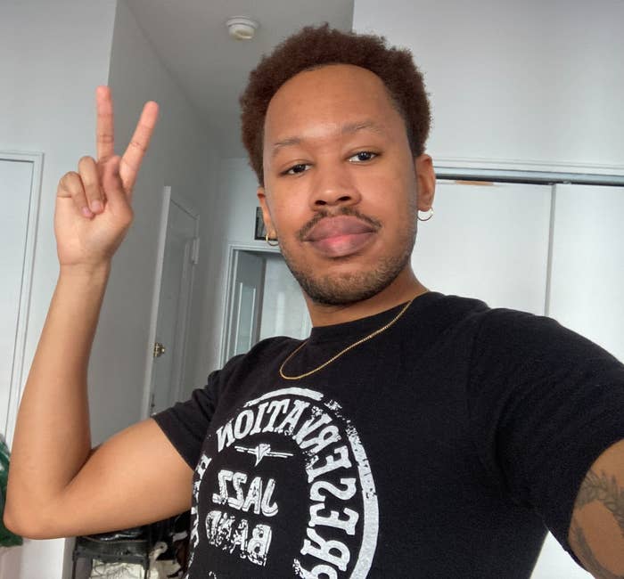A person with short curly hair wearing a black graphic T-shirt with "Reservation Jazz Band" on it, posing indoors while making a peace sign