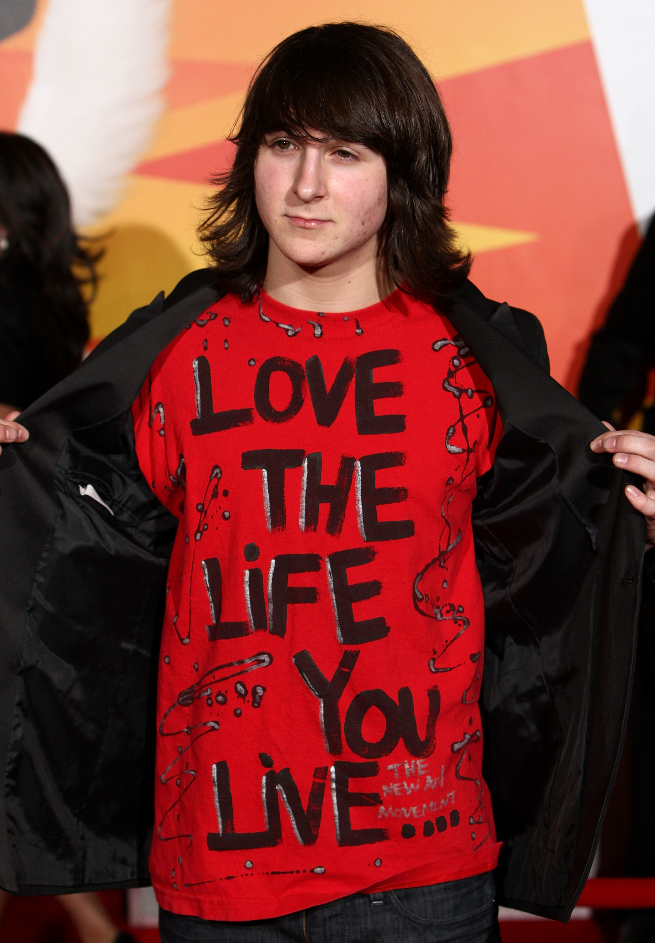 Mitchel Musso in a red T-shirt that reads "LOVE THE LIFE YOU LIVE" under an open black jacket, posing at an event