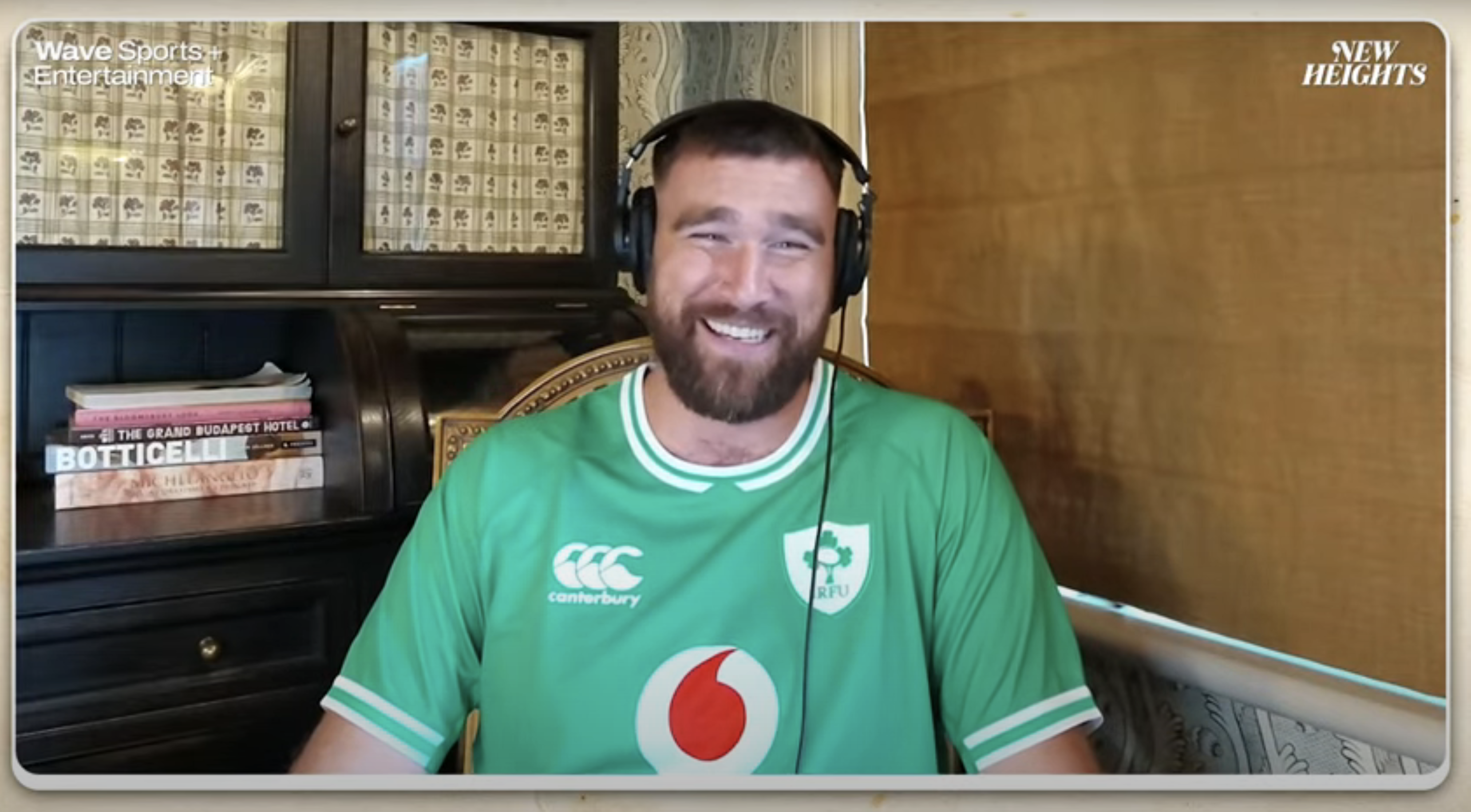 Travis Kelce is smiling, wearing a green sports jersey with headphones on, during a virtual interview with 'New Heights' branding in top corners