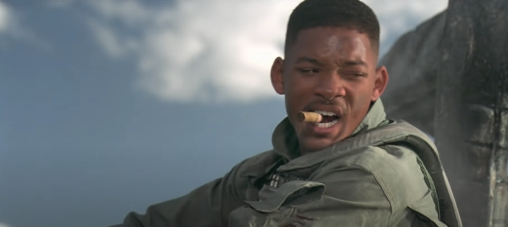 Will Smith wearing a flight suit and holding a cigar while looking off to the side