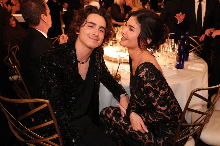 Timothée Chalamet and Kylie Jenner seated together at a formal event, both dressed in elegant black attire and smiling