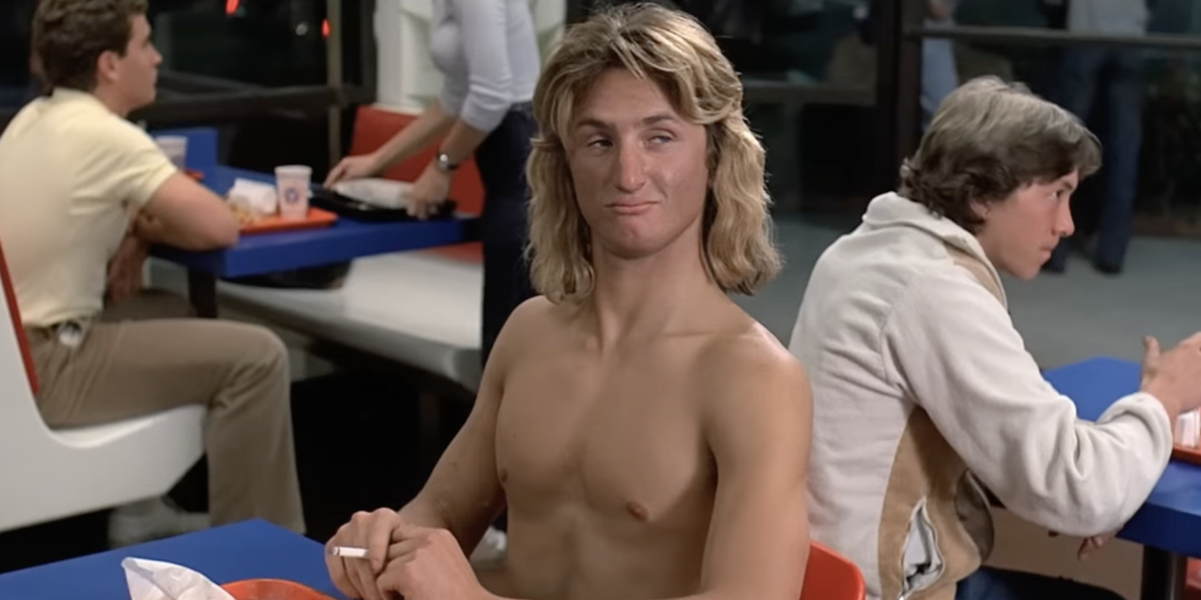 Shirtless Sean Penn, with long hair, sits at a fast-food restaurant table, holding a cigarette, while other patrons dine in the background