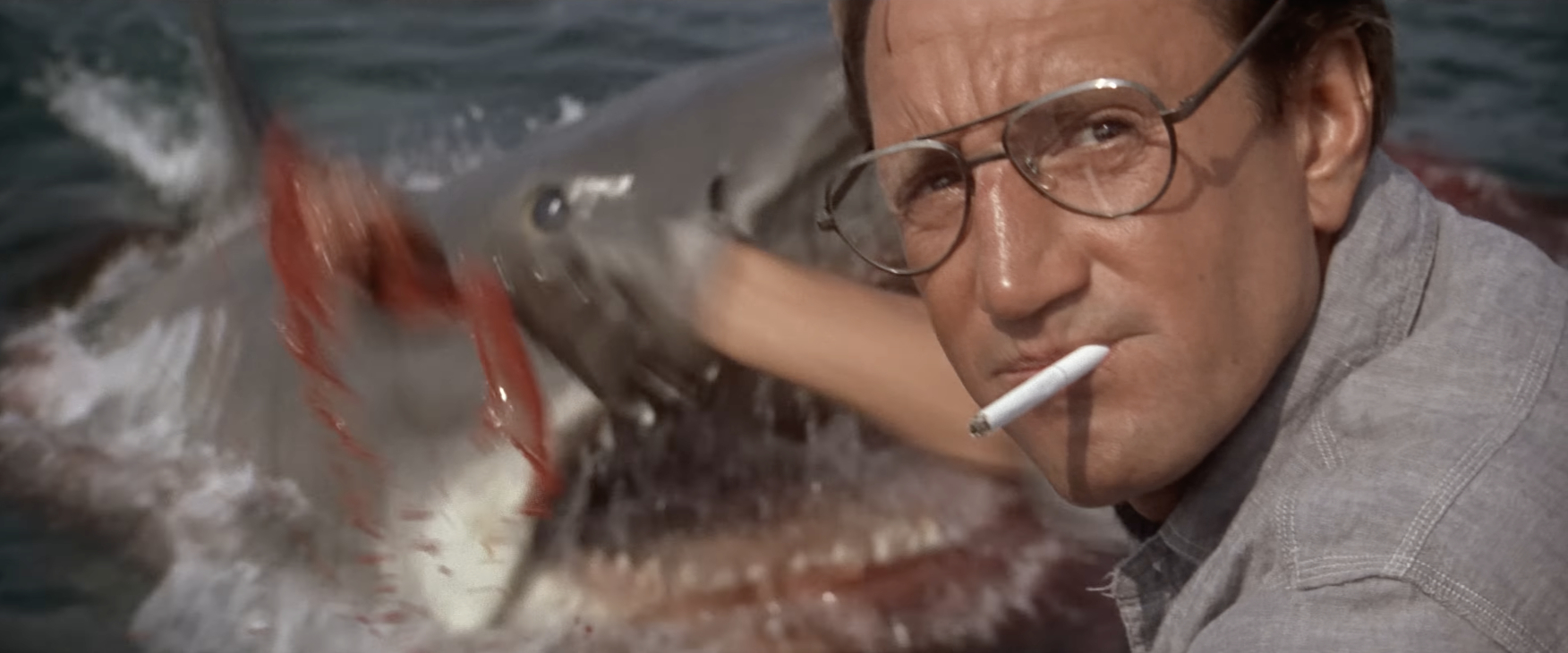 A man with glasses and a cigarette in his mouth stands in front of a large shark in the ocean
