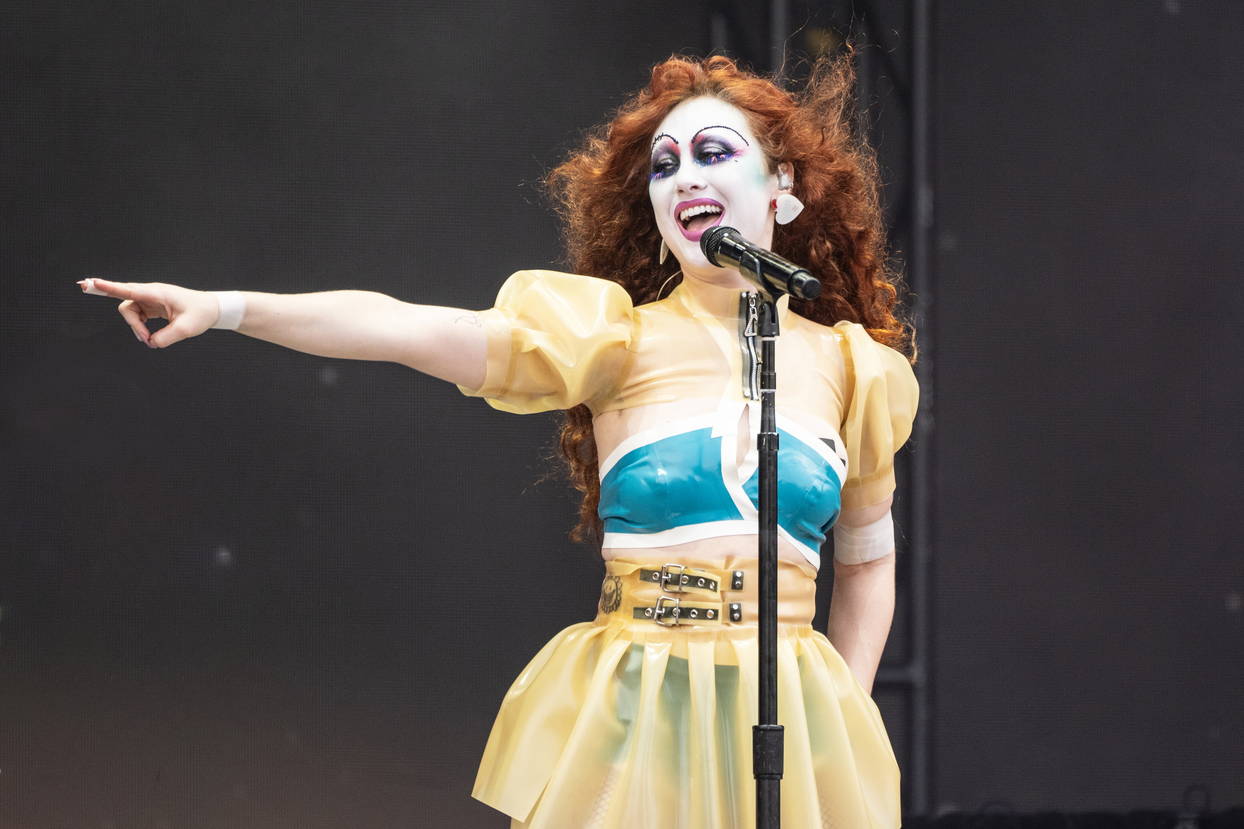 Chappell Roan performing on stage with dramatic makeup, curly hair, and a colorful dress, pointing to the side