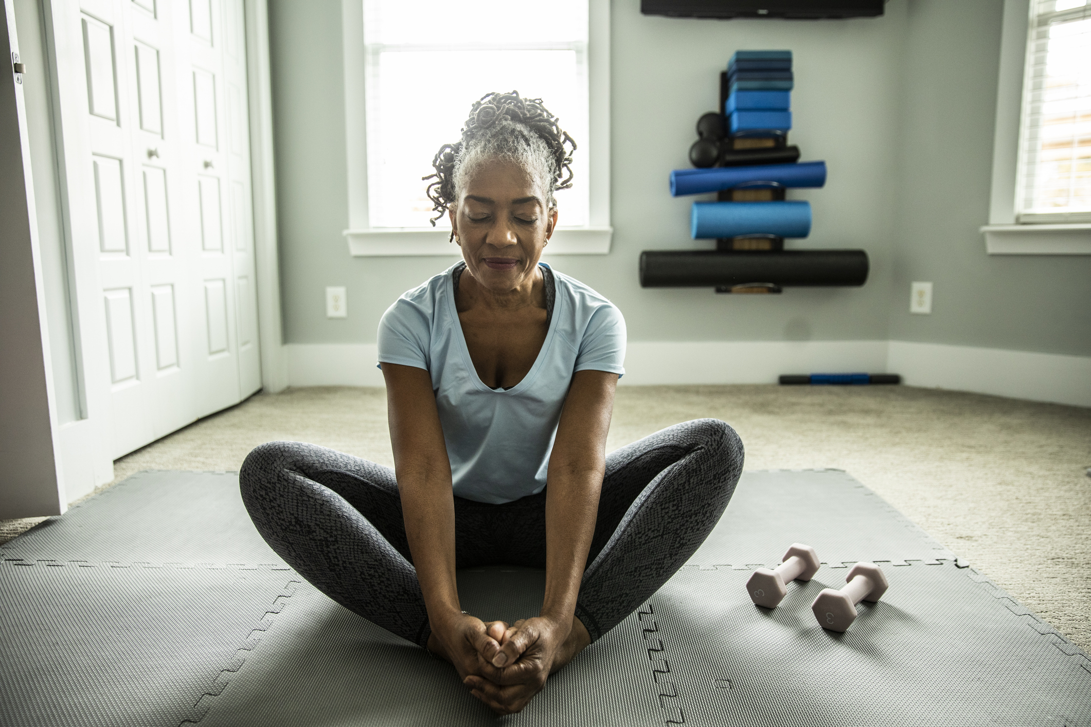 A woman dressed in athletic wear sits on a mat in a yoga position with dumbbells beside her, appearing calm and focused in a home gym setting