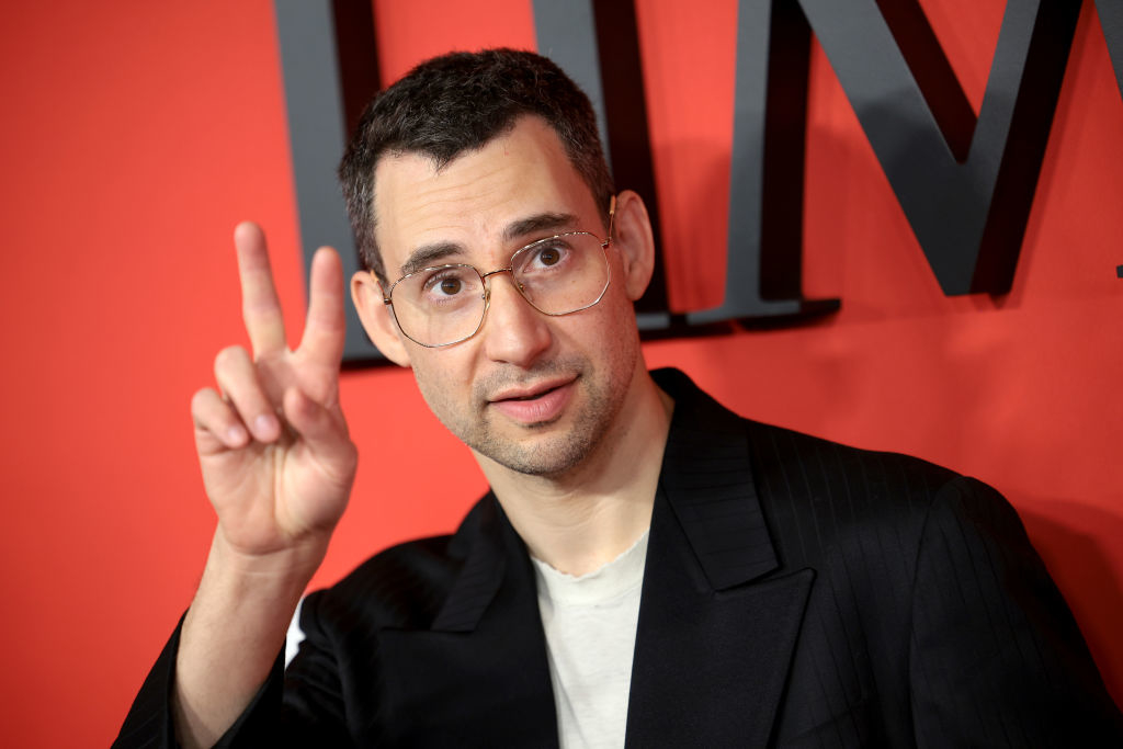 A person with short hair and glasses wearing a blazer over a light shirt poses in front of a red background, making a peace sign with their hand