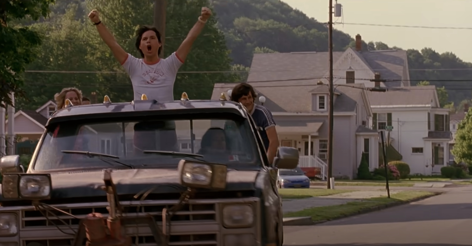 Group of people in a car with a woman standing through the sunroof, raising her arms and cheering in a suburban neighborhood