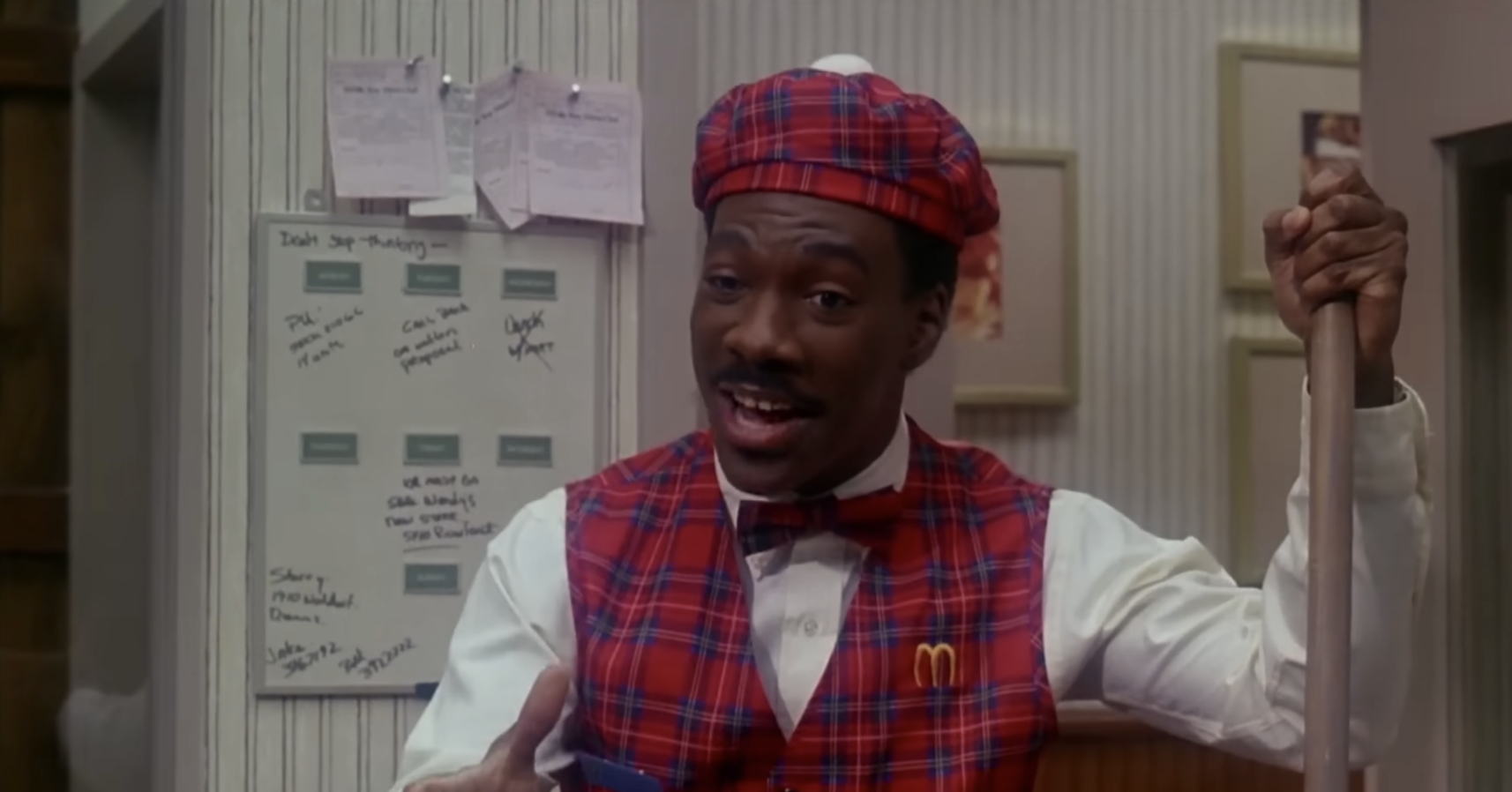 Eddie Murphy dressed as a McDonald's employee in a red plaid uniform with a hat, portrayed in a scene where he is cleaning while talking. Notes are pinned on a board behind him