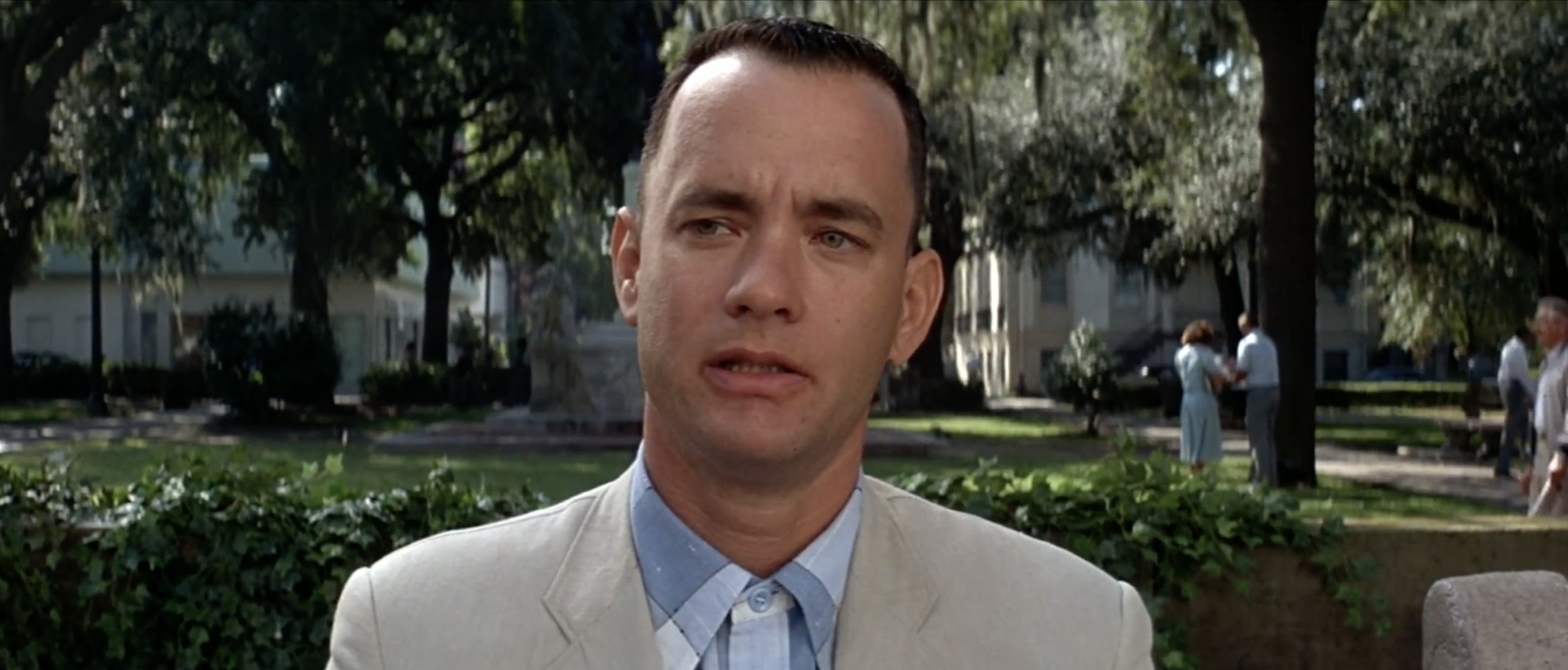 Tom Hanks wearing a light-colored suit, sitting on a bench in a park with a thoughtful expression. Trees and people are visible in the background