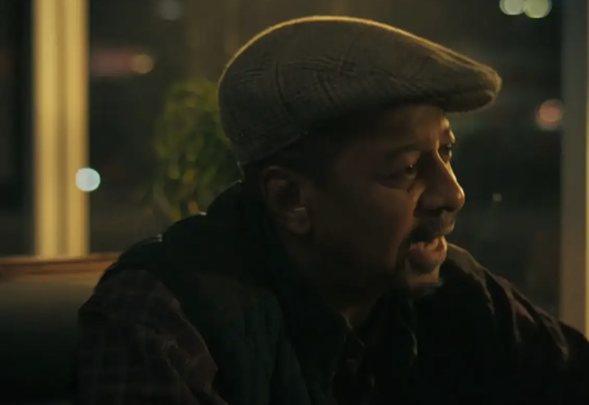 A man in a flat cap and patterned shirt sits indoors at a table in a dimly lit setting, speaking