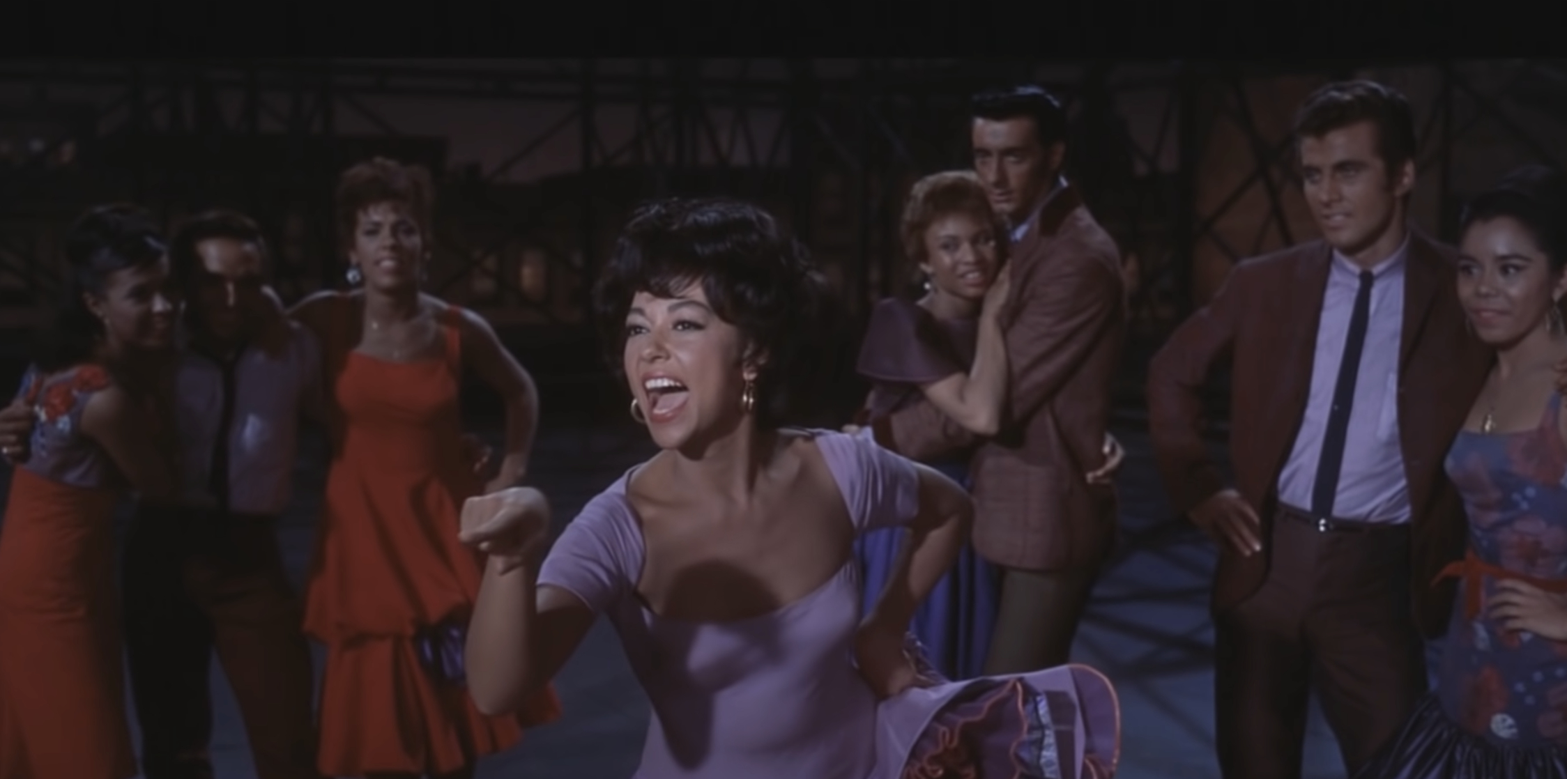 Group scene from "West Side Story": Rita Moreno in the foreground, dancing energetically with her co-stars in 1960s-style clothing in an urban setting