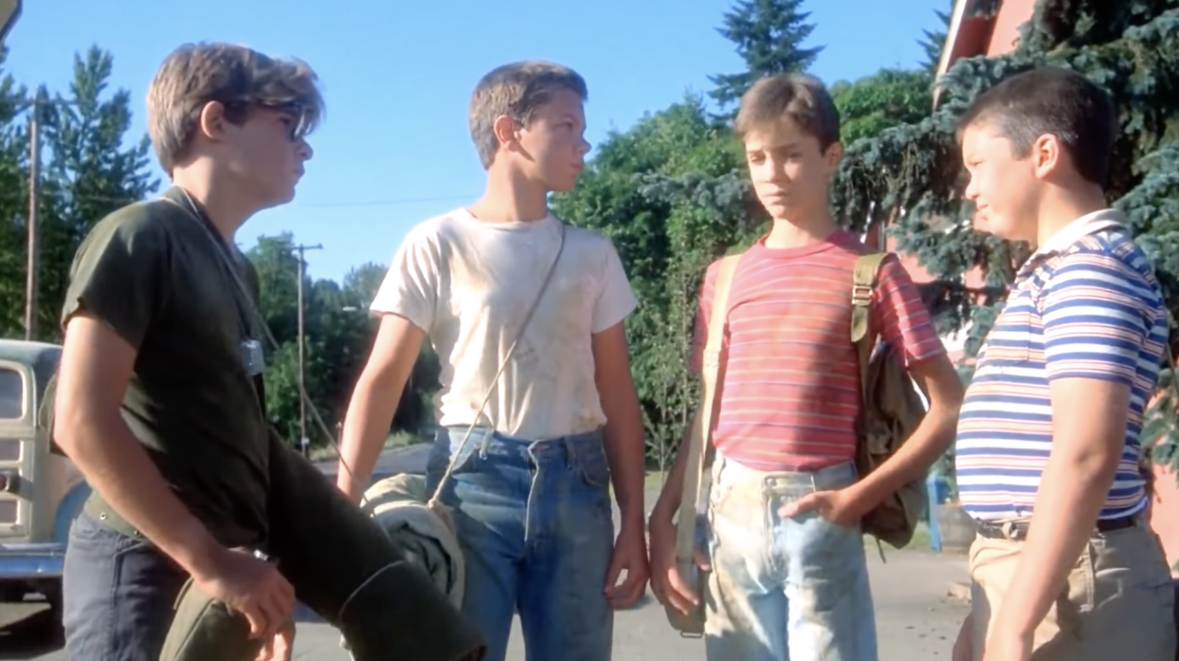 Four boys, including Wil Wheaton and River Phoenix, are standing outdoors talking to each other. They are casually dressed in T-shirts and jeans